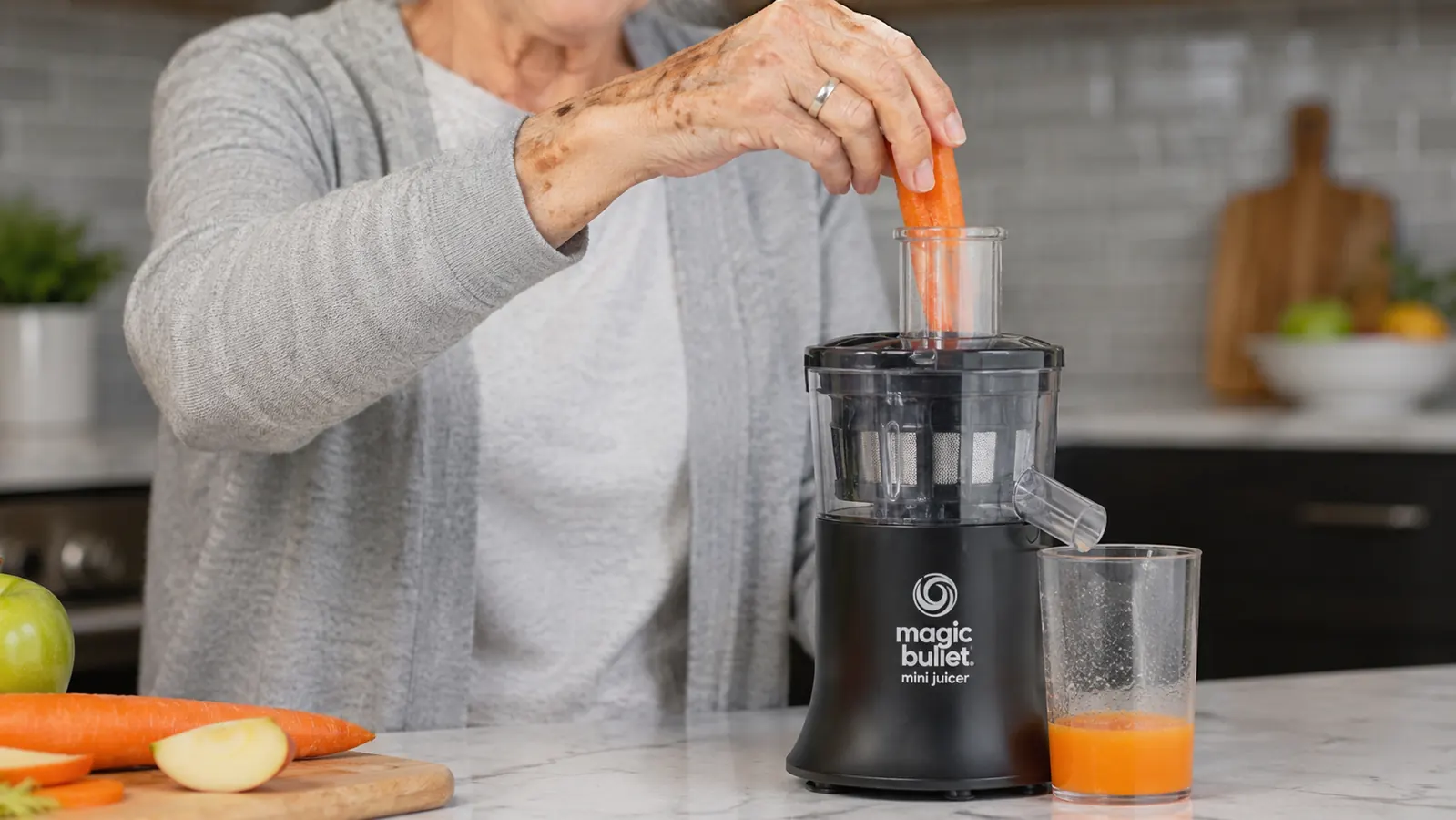 An older woman's hands feeding a carrot piece into the narrow feed chute of a small black countertop juicer